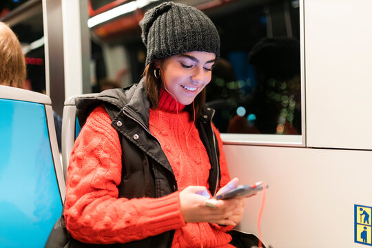 Smiling young woman using smart phone and listening music through in-ear headphones in tram