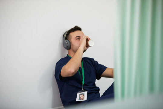 Male Nurse With Headphones Drinking Coffee By Wall In Medical Room