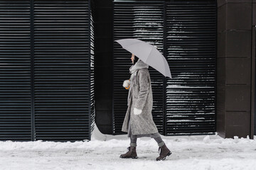 Contemplative woman with umbrella walking on footpath by wall at winter