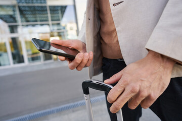 Hands of man holding smartphone and suitcase handle
