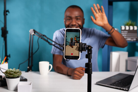 Closeup of live vlog setup with smarthone on stand filming vlogger waving hello sitting at desk with professional microphone. Selective focus on mobile phone recording social media influencer.