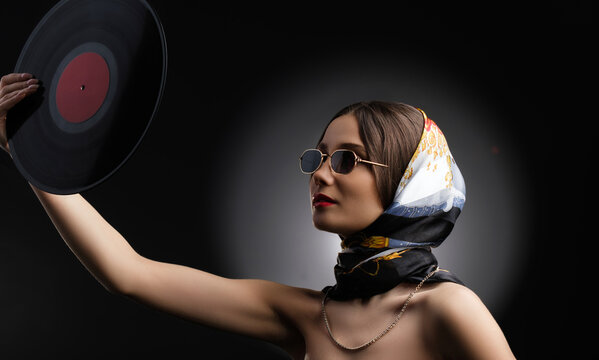 A photo of glamorous attractive young woman in sunglasses and beautiful shawl on a head holding vinyl LP in outstretched hand.