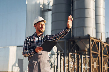 In grey clothes. Construction worker in uniform is outdoors near the factory
