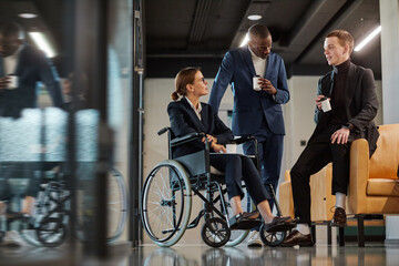 Fototapeta na wymiar Full length portrait of diverse business team with young woman in wheelchair chatting to male colleagues in modern office
