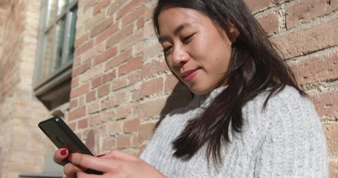 Portrait of young woman smiling and text messaging on street against brick wall