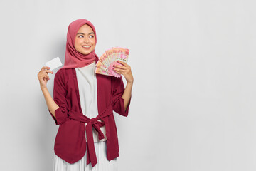 Cheerful beautiful Asian woman in casual shirt and hijab holding Indonesian rupiah banknotes, showing credit card isolated over white background. People religious lifestyle concept