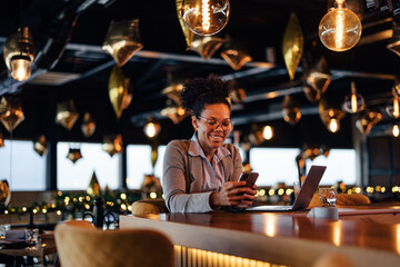 Smiling African-American woman, relaxing at the bar counter, using her phone.