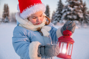 A Christmas child on the street. A girl in winter in a blue jacket and a red hat holds a red lantern candlestick in her hands and looks at it.