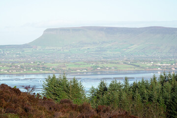 Obraz premium Green forest in foreground, Benbulben flat top mountain in the background. Blue cloudy sky. Irish nature. County Sligo, Ireland