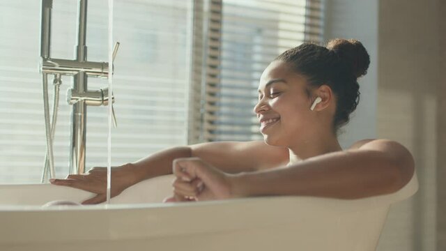 Young african american lady enjoying music in wireless earphones, resting in hot bathtub with pouring water, zoom in