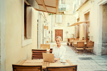 Technology and travel. Working outdoors. Freelance concept. Pretty young woman using laptop in sidewalk cafe on ancient europian street.