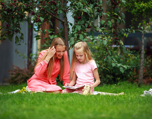 Naklejka premium Mother and daughter on green grass lawn reading book together in backyard garden