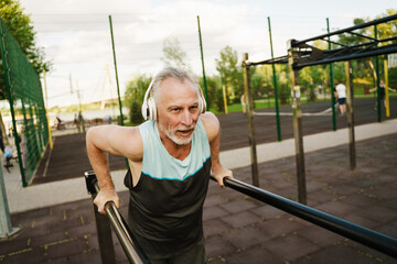 Grey senior sportsman listening music while working out on parallel bars