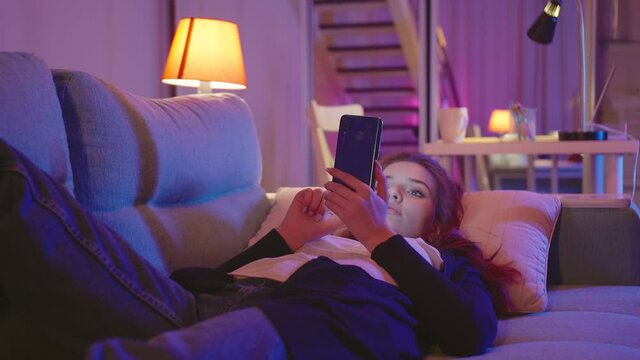 Relaxed Young Woman Using Smart Phone Surfing Social Media, Checking News, Playing Mobile Games Or Texting Messages Laying On Sofa. Millennial Lady Spending Time At Home With Cell Gadget Technology