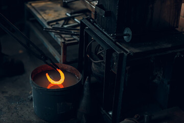 Fototapeta na wymiar Blacksmith forges and tempering metal horseshoe in jar with water at forge.
