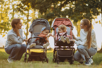 Two women friends walking with baby prams and their kids in park