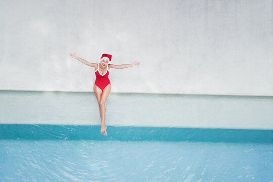 Enjoying christmas holidays and vacation. Top view of excited young woman in red swimsuit  and  santa claus hat rising hands up near swimming pool.