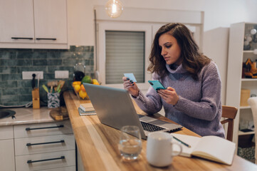 Hispanic woman with laptop using credit card and smartphone in kitchen at home