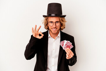 Young magician caucasian man holding magic cards isolated on white background cheerful and confident showing ok gesture.