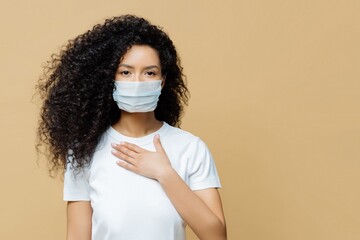 Serious Afro American woman wears medical face mask, has problems with breathing, presses hand to...