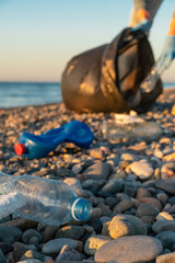 Fototapeta premium Close-up of plastic bottles against the background of a young woman with a black garbage bag on a pebble beach of the sea at sunset.Concept of ecology,volunteering.Vertical photo