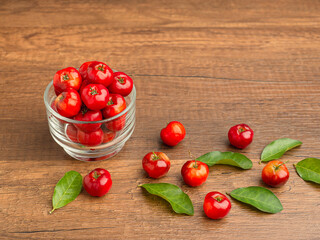 Ripe red acerola cherries in glass and green leaves scattered on a wooden table.