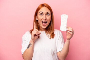 Young caucasian woman holding sanitary napkin isolated on pink background pointing upside with opened mouth.