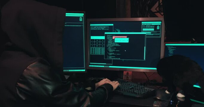 Hacker in a hood sits in front of computer screens and hacks databases. The concept of cyberterrorism, server hacking and cyberattacks. There is a black cat next to the man