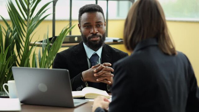 Young businessman talking with woman and sitting at table with laptop in company spbi. Closeup view of American African man talks to female colleague and looks with smile, discusses work and sits at