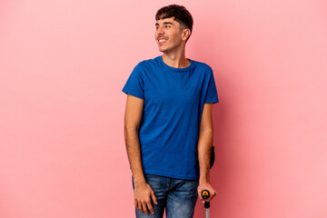 Young mixed race man with crutches isolated on pink background looks aside smiling, cheerful and pleasant.