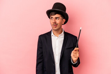 Young caucasian wizard man holding a wand isolated on pink background confused, feels doubtful and...