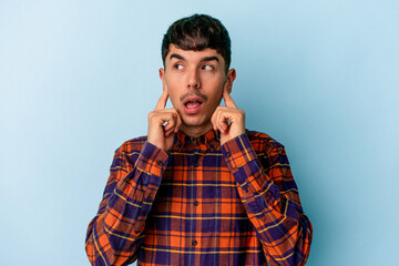 Young mixed race man isolated on blue background covering ears with fingers, stressed and desperate by a loudly ambient.