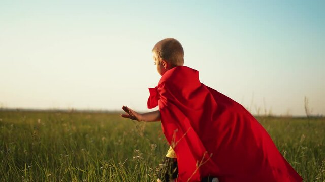 Happy kid in superhero costume. Boy leader run on green grass in park. Childhood dream, child is winner. Boy in red superhero costume in rays of sun. Child hero run on green grass. Child dream in park