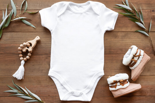 Mockup of white baby bodysuit on dark wood background with greenery, booties and toy. Blank baby clothes template in farmhouse style, flat lay.