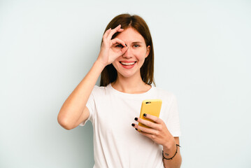 Fototapeta premium Young English woman holding mobile phone isolated on blue background excited keeping ok gesture on eye.