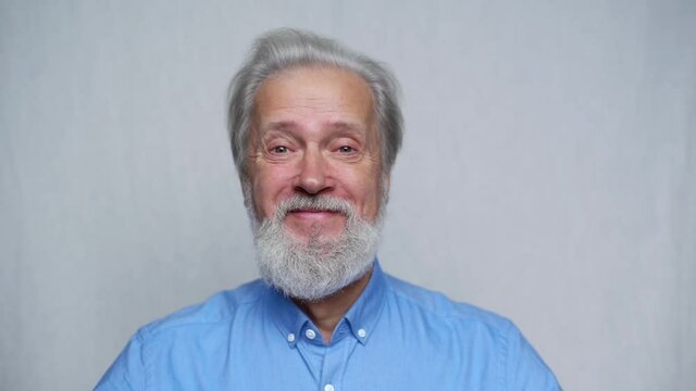 Close-up studio portrait of cheerful gray-haired mature man smiling looking at camera on white isolated background. Front view of positive bearded old aged gentleman posing in studio, slow motion.