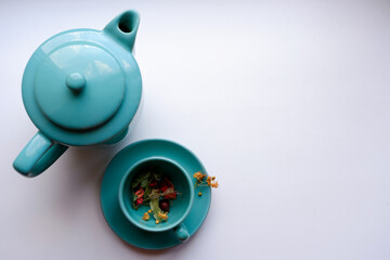 Top view: a mug on a saucer with colored herbs for tea and a teapot on a white background
