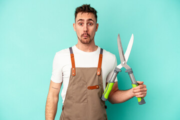 Young gardener caucasian man holding a scissors isolated on blue background shrugs shoulders and...
