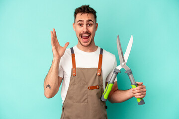 Young gardener caucasian man holding a scissors isolated on blue background receiving a pleasant surprise, excited and raising hands.