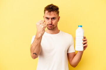 Young caucasian man holding a bottle of mil isolated on yellow background showing fist to camera, aggressive facial expression.