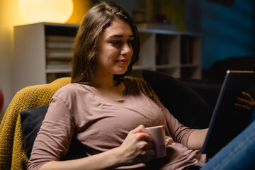 young woman relaxing on sofa at home. she is using digital tablet. night evening scene