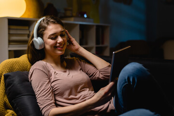 Obraz premium young woman relaxing on sofa at home. she is using digital tablet and listening music on headphones . night evening scene