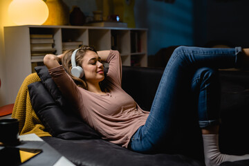 young woman relaxing on sofa at home. she is listening music on headphones . night evening scene