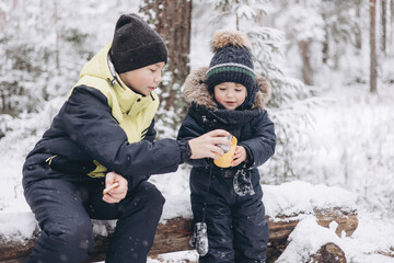 Fototapeta na wymiar Happy teenage boy and todler drinking tea from thermos and talking sitting together on log in winter snowy forest. Hot beverage in cold weather. Children having picnic in winter season outdoors