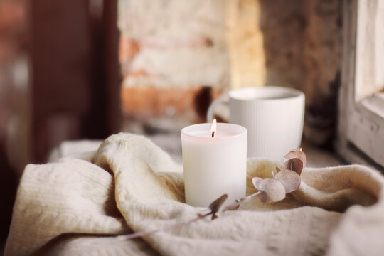 White burning candle on woolen plaid and cup of tea on windowsill at home in cozy atmosphere