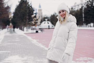 Beautiful girl in fashionable winter clothes (white down jacket, stylish knitted hat, mittens)...