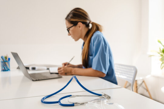 White woman doctor writing down notes while working with laptop
