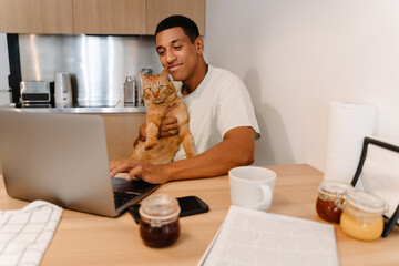 Black man holding his cat while working with laptop and having breakfast