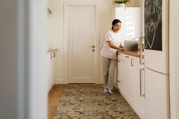 Young hispanic woman smiling while using laptop in kitchen