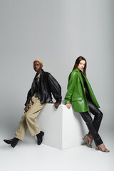 full length view of young and stylish multiethnic couple posing on white cube on grey.